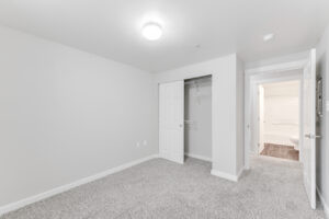 Interior Unit Bedroom, White walls, neutral toned carpeting, wood like floors in bathroom, ceiling light fixture, sliding closet door.