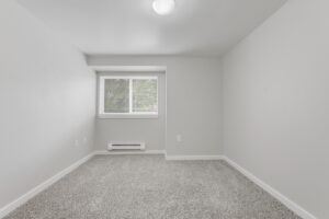 Interior Unit Bedroom, central AC, window in left hand corner, neutral toned carpeting, white walls.
