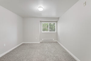 Interior Unit Bedroom, central AC, window in left hand corner, neutral toned carpeting, white walls.