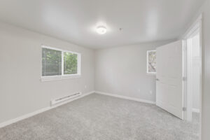 Interior Unit Bedroom, white walls central ac, windows on two walls, ceiling light fixture, neutral toned carpeting.