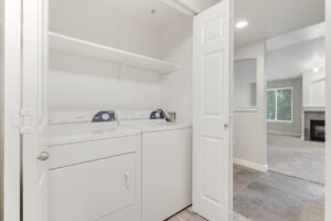 Interior Unit Linen Closet, washer and dryer, storage shelf above laundry machines, white walls, neutral toned carpeting, tile floor in nearby foyer.