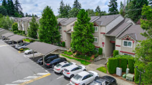 Aerial Exterior of residential buildings, covered and non covered parking, parking out front, meticulous landscaping around building, trees planted along sidewalks, white buildings with red trim.