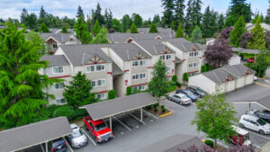 Aerial Exterior residential building, white building with red trim, meticulous landscaping, covered and uncovered parking, lush foliage, dense woods behind building.