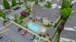 Aerial Exterior of leasing office and community swimming pool, meticulous landscaping, parking in front, covered and uncovered parking, lush foliage, hot tub, bistro tables with umbrellas.